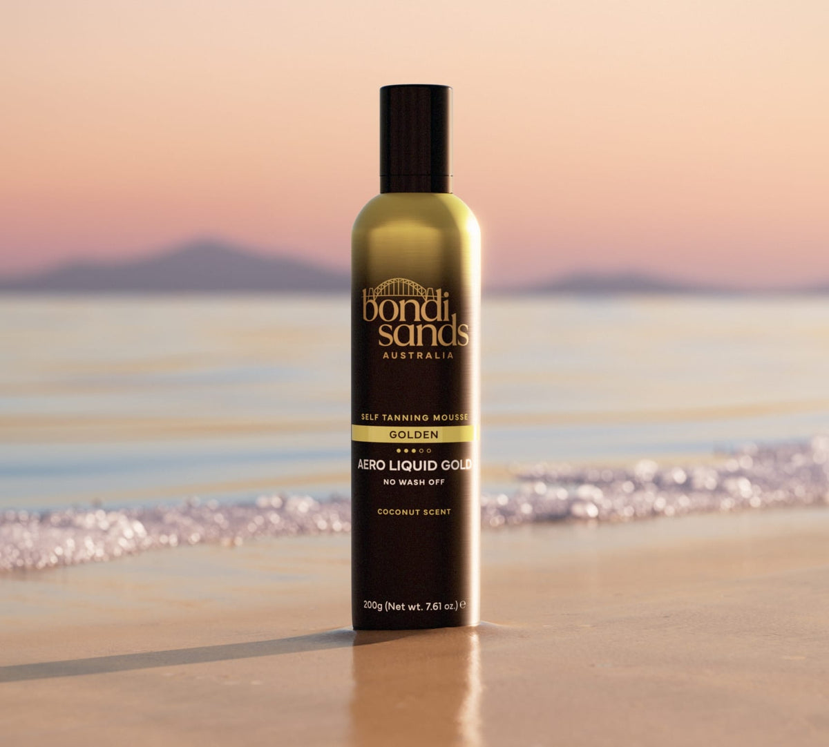 A bottle of BondiSandsEU Liquid Gold Aero Self Tanning Mousse stands upright on wet beach sand, with calm water and distant mountains in soft evening light.