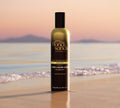 A bottle of BondiSandsEU Liquid Gold Aero Self Tanning Mousse stands upright on wet beach sand, with calm water and distant mountains in soft evening light.