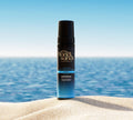 A BondiSandsEU Express Self Tanning Foam bottle stands upright on a sandy beach, offering a customisable tan with the tranquil blue ocean and clear sky in the background.