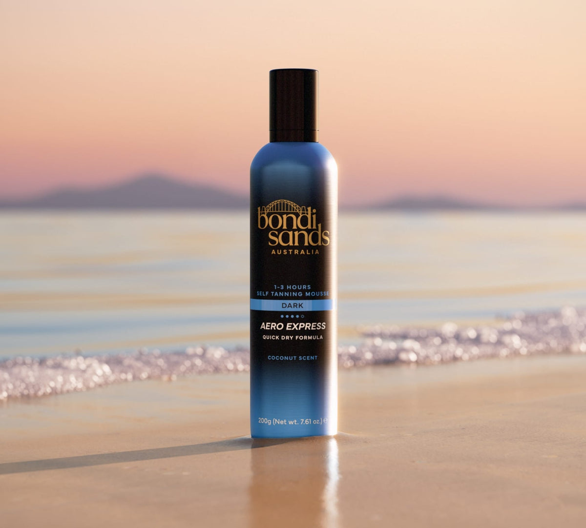 A bottle of BondiSandsEU Express Aero Self Tanning Mousse stands upright on wet beach sand, offering a quick-dry, deep bronze tan as gentle waves and distant mountains enhance the sunset view.