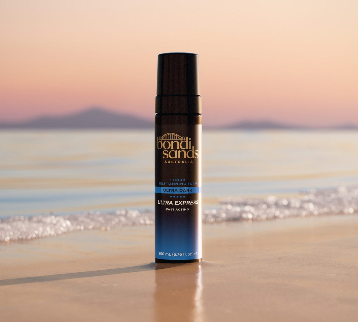 Bondi Sands Europe Ultra Express Self Tanning Foam stands upright on wet sand by the shoreline, with gentle waves, calm water, and distant mountains at sunset—ideal for fast tanning results.