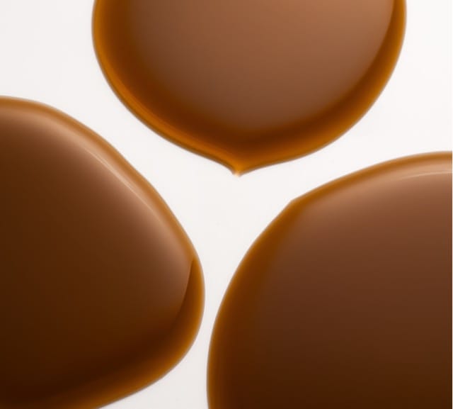 Three overlapping puddles of BondiSandsEU Liquid Gold Self Tanning Oil are spread on a white surface, forming smooth, rounded shapes with a rich brown hue and soft reflections.