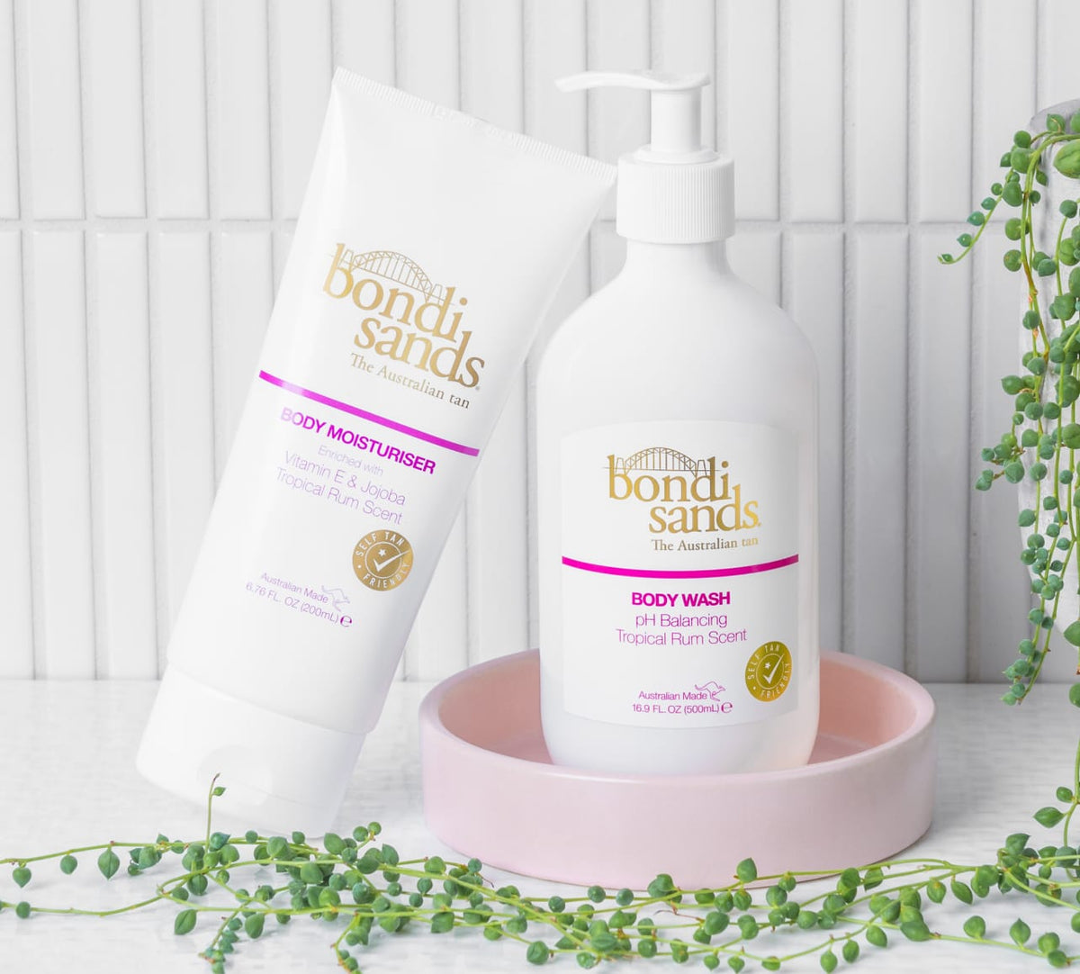 The BondiSandsEU Tropical Rum Body Wash sits on a white surface with a pink dish and green plants, set against crisp white tiles.