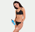 A woman in a black bikini smiles at the camera as she applies BondiSandsEU Aero Dark Self Tanning Mousse Bundle to her thigh, holding its blue bottle in her left hand against a plain light background.