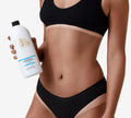 A woman in black lingerie holds a bottle of BondiSandsEU Light-Medium Salon Solution 1L Duo, revealing defined tan lines and a bronzed glow, on a plain white background.