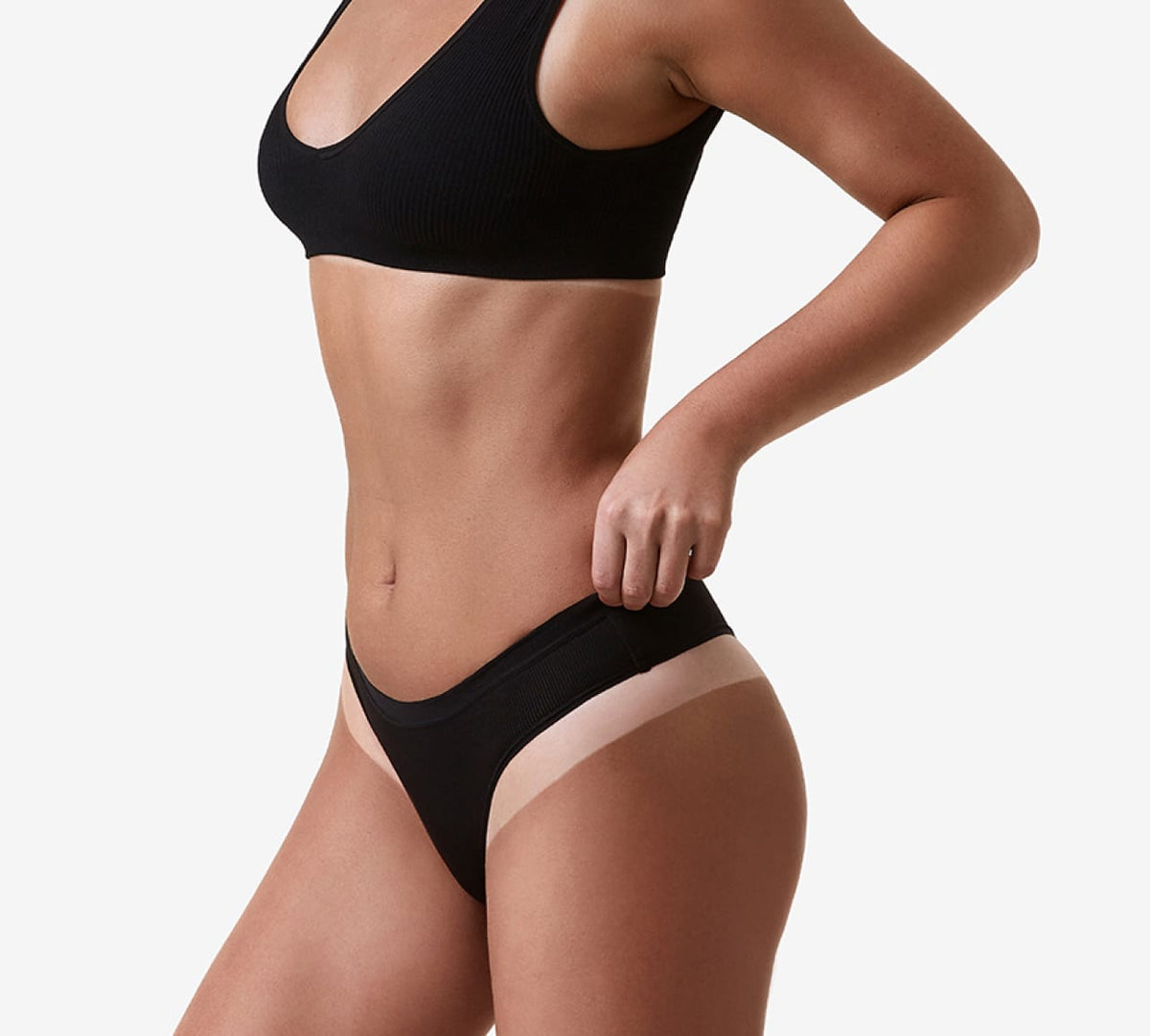 A woman with a bronzed glow, achieved using BondiSandsEU Light-Medium Salon Solution 1L Duo, stands against a white background in black lingerie with beige trim, her left hand on her hip and her torso shown from shoulders to upper thigh.