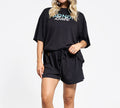 Model wears BondiSandsEU Bondi Babe Short Tanning PJs: a loose black t-shirt with “Bondi Babe” text and matching drawstring shorts, perfect as loungewear or self-tan pyjamas. Her face is partially out of frame against a plain white background.