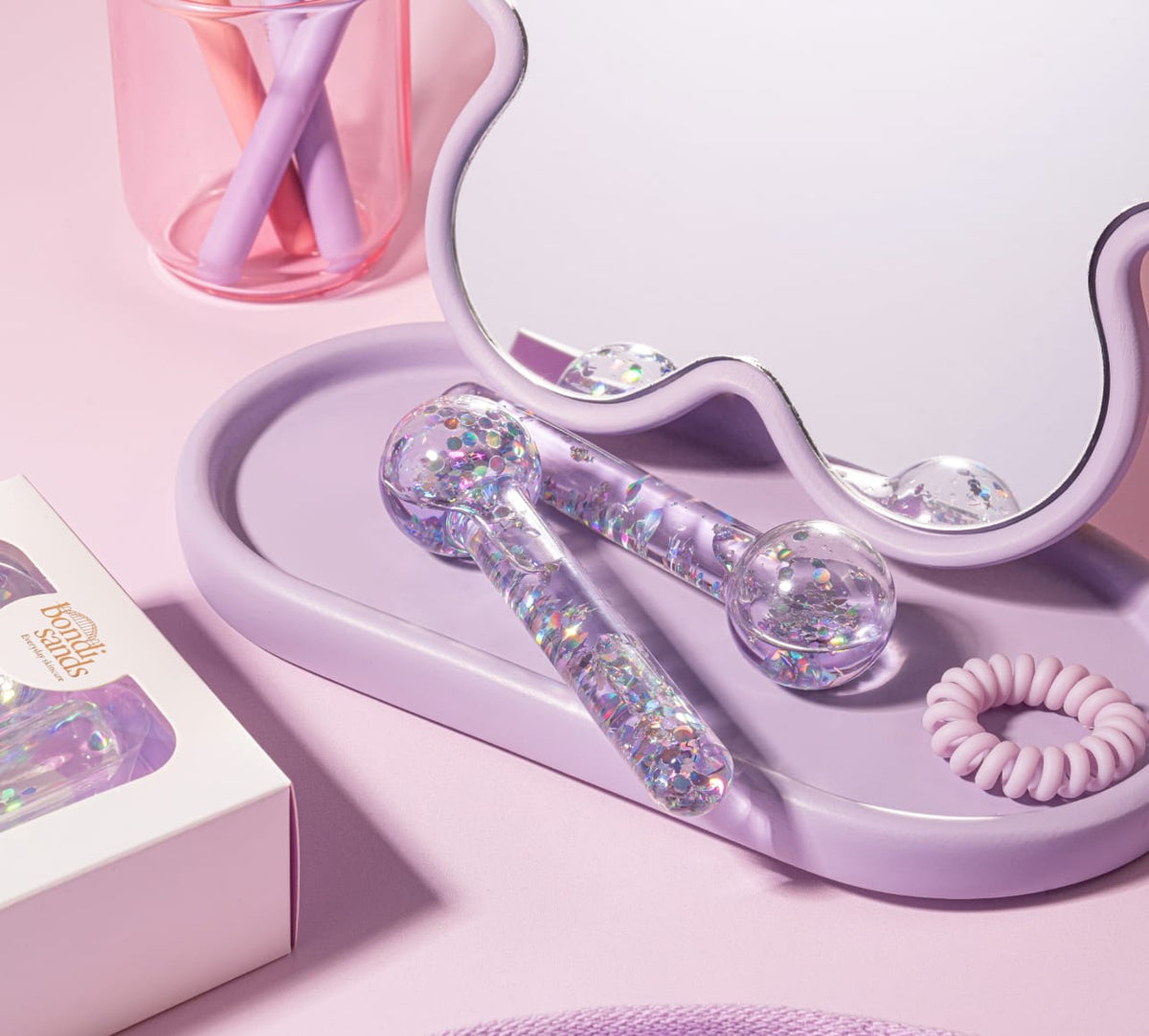 Two sparkly, clear De Puff Cooling Facial Globes by BondiSandsEU rest on a lavender tray near a wavy mirror, with a pink spiral hair tie, makeup brushes in a pink cup, and a Benefits Smile box—ideal for de-puffing and lymphatic drainage.