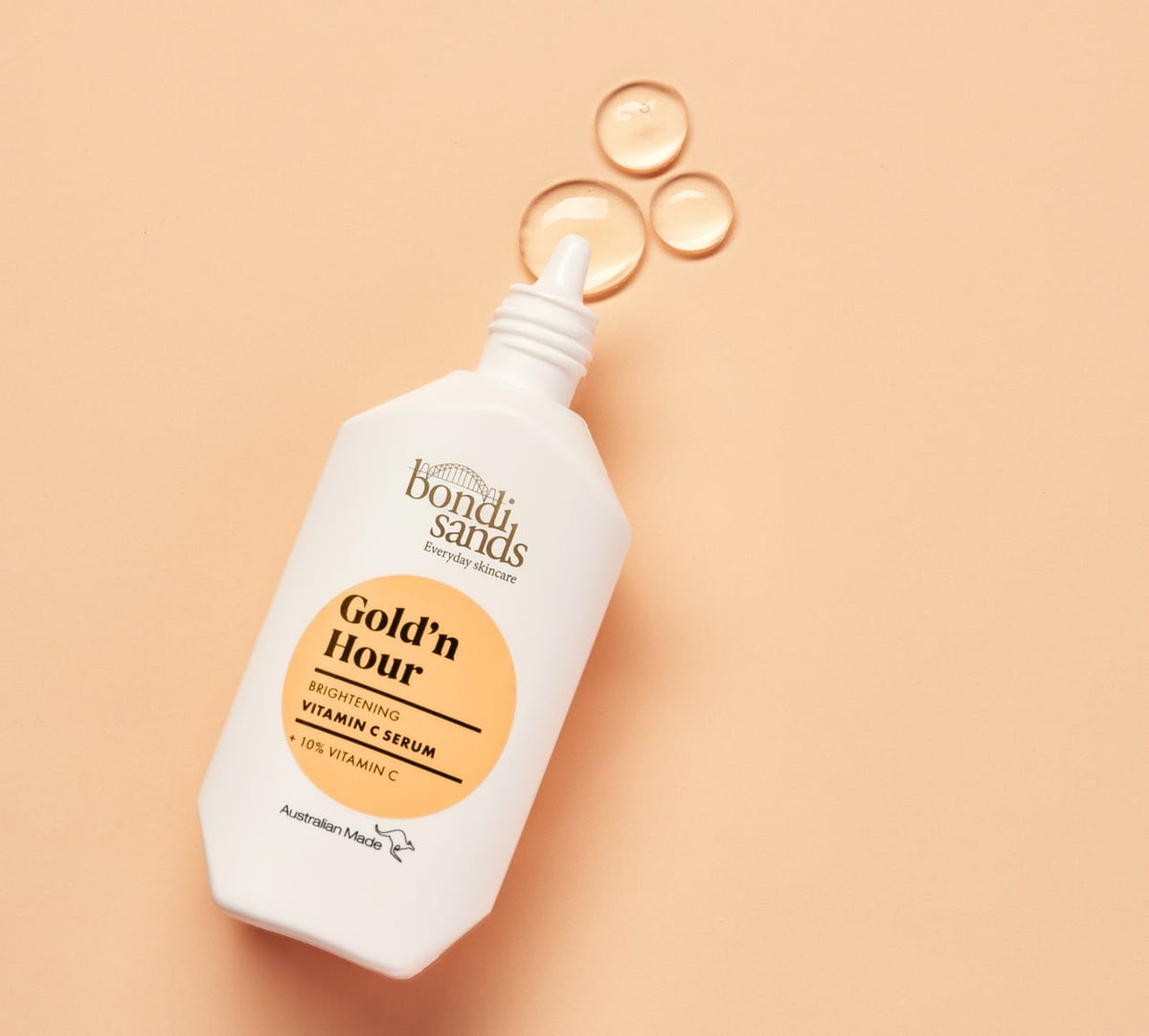 A white BondiSandsEU Gold'n Hour Vitamin C Serum bottle with a pointed tip rests on a peach surface, displaying three clear drops—ideal for brightening skin and enhancing radiance.