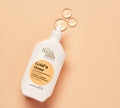 A white BondiSandsEU Gold'n Hour Vitamin C Serum bottle with a pointed tip rests on a peach surface, displaying three clear drops—ideal for brightening skin and enhancing radiance.