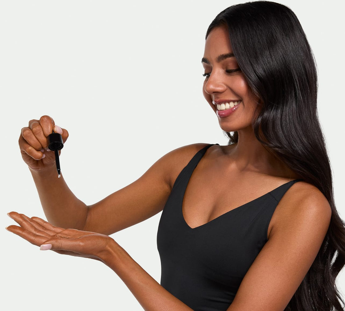 A woman with long, dark hair in a black sleeveless top smiles, holding a dropper above her palm as she prepares to apply BondiSandsEU Dark Self Tanning Face Drops for a radiant, bronzed look. The background is light and uncluttered.