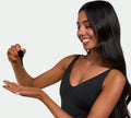 A woman with long, dark hair in a black sleeveless top smiles, holding a dropper above her palm as she prepares to apply BondiSandsEU Dark Self Tanning Face Drops for a radiant, bronzed look. The background is light and uncluttered.