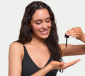 A woman with long, wavy brown hair and bronzed skin smiles while using BondiSandsEU Light-Medium Self Tanning Face Drops, dispensing the non-comedogenic formula into her palm against a simple light background.