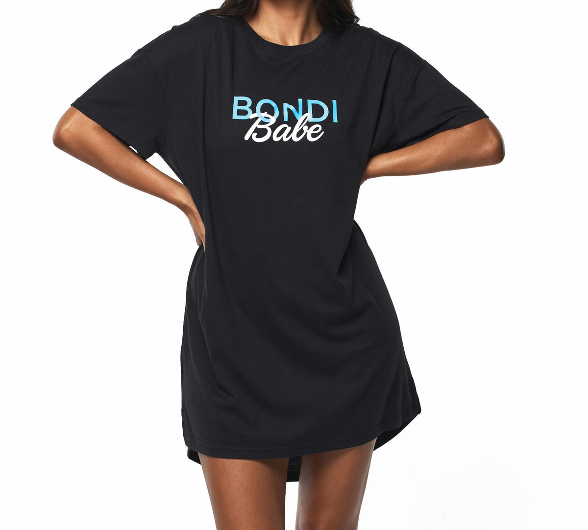 A person wears the BondiSandsEU Bondi Babe Self Tanning Tee—black, loose-fitting, and featuring "BONDI Babe" in blue and white text—for a comfy sleep look against a white background.