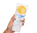 A hand holds BondiSandsEU SPF 50+ Everyday Body Sunscreen Lotion—a white tube with a yellow sunset logo and blue text, offering very high protection, no added fragrance, and 72-hour hydration.