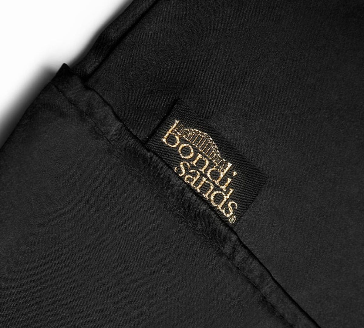 A close-up of black fabric features a stitched black label reading "BondiSandsEU" in gold. The Self Tanning Pillow Protector is sewn onto a folded corner, ensuring mess-free tanning and preventing tan transfer.