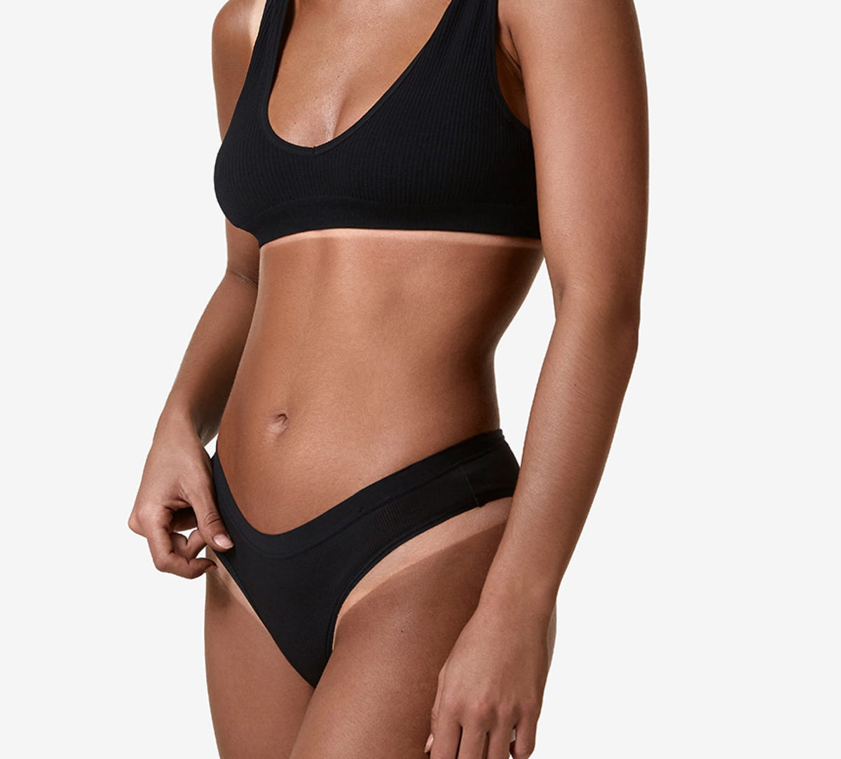 A woman in a black ribbed sports bra and matching underwear stands against a white background, highlighting her flawless tan—ideal for fans of the BondiSandsEU Salon Solution Ultra Dark. Her head and legs are not visible.