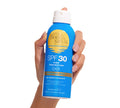 A hand holds a blue can of BondiSandsEU SPF 30 Everyday Sunscreen Aerosol Mist with yellow and white text and a blue cap, set against a plain white background.