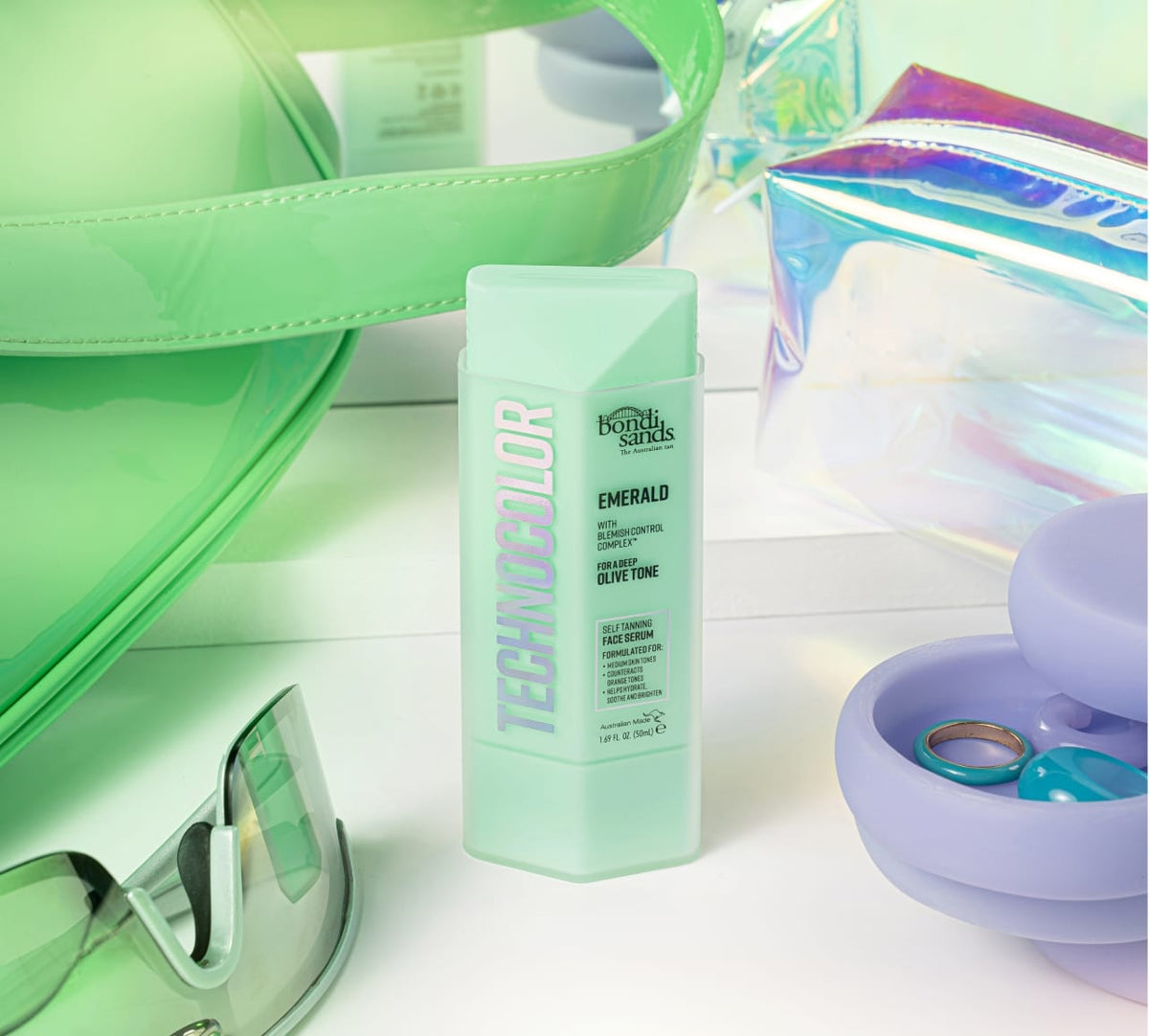 A mint green bottle of BondiSandsEU Emerald Technocolor Self Tanning Face Serum stands upright among pastel accessories like sunglasses, bags, and a purple contact lens case with lenses inside.