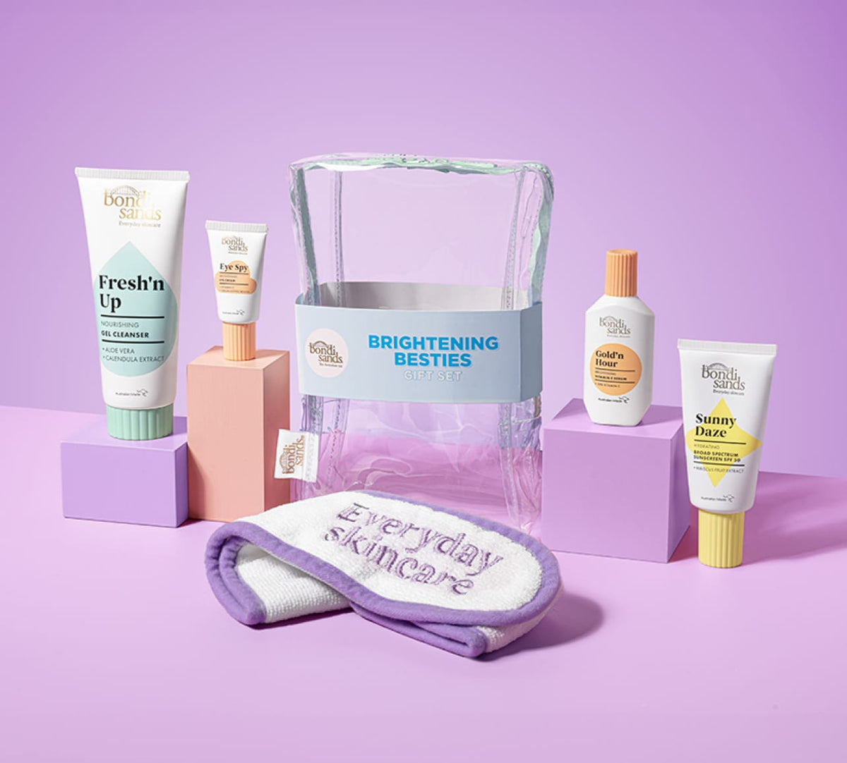 The BondiSandsEU Brightening Besties Gift Set features Vitamin C serum, cleansers, sunscreen, a "Brightening Besties" cosmetic bag, and an "Everyday Skincare" headband, all displayed on a pink surface.