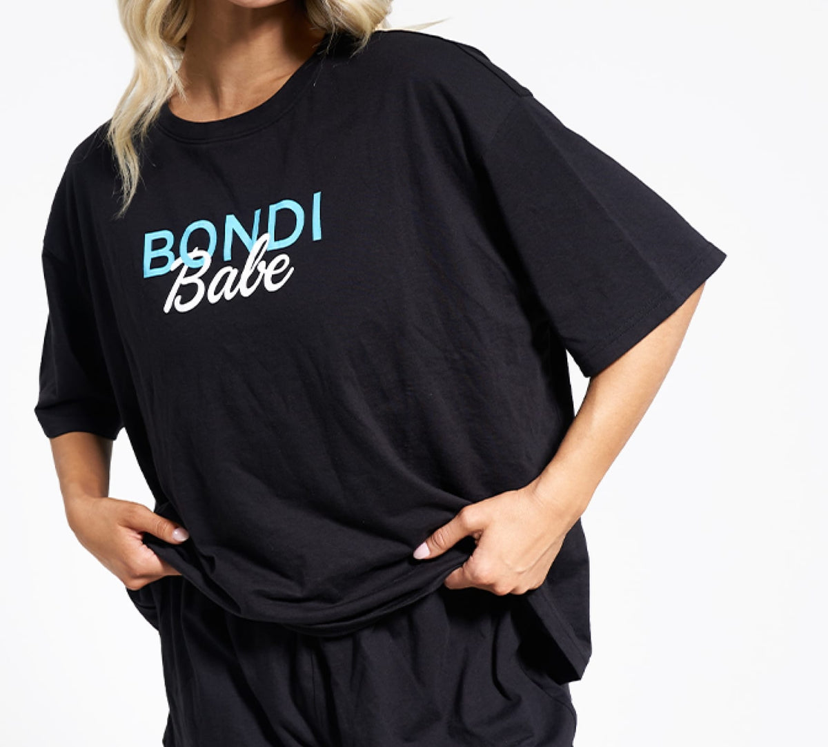 A person gently holds the hem of a loose black BondiSandsEU t-shirt with “Bondi Babe” in blue and white, perfect for relaxing or pairing with the Bondi Babe Short Tanning PJs. Their head is partially out of frame.