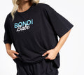 A person gently holds the hem of a loose black BondiSandsEU t-shirt with “Bondi Babe” in blue and white, perfect for relaxing or pairing with the Bondi Babe Short Tanning PJs. Their head is partially out of frame.