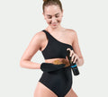 A woman in a black one-shoulder swimsuit smiles while pumping self-tanning mousse onto the BondiSandsEU Application Mitt, ready to achieve a flawless, streak-free glow.