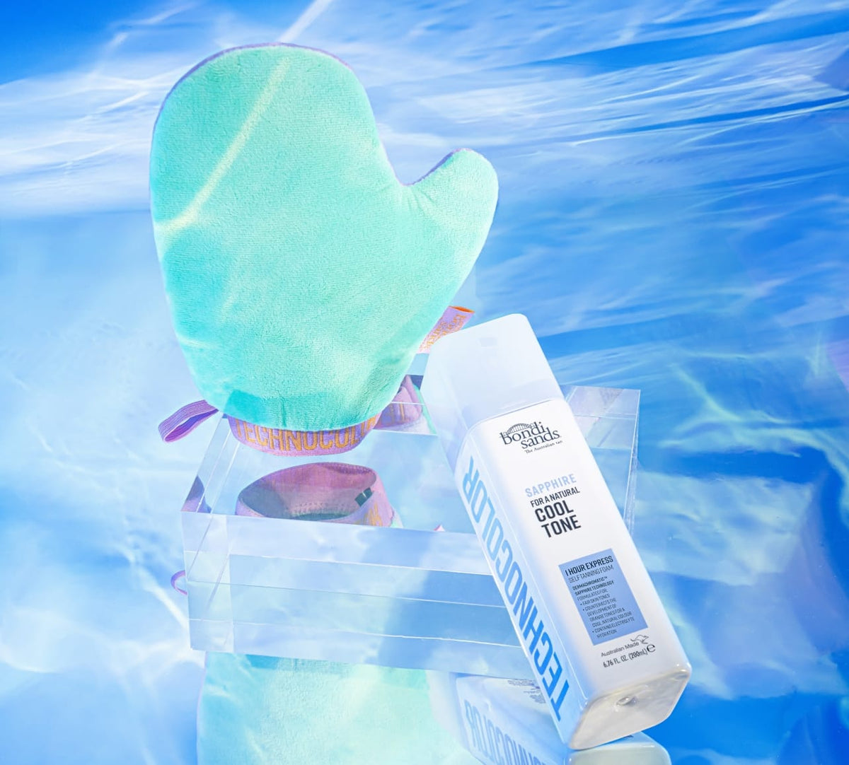 A turquoise tanning mitt and a bottle of BondiSandsEU Sapphire Technocolor 1 Hour Express Self Tanning Foam sit on a clear acrylic tray, reflected against a blue watery background for an effortless, natural glow.