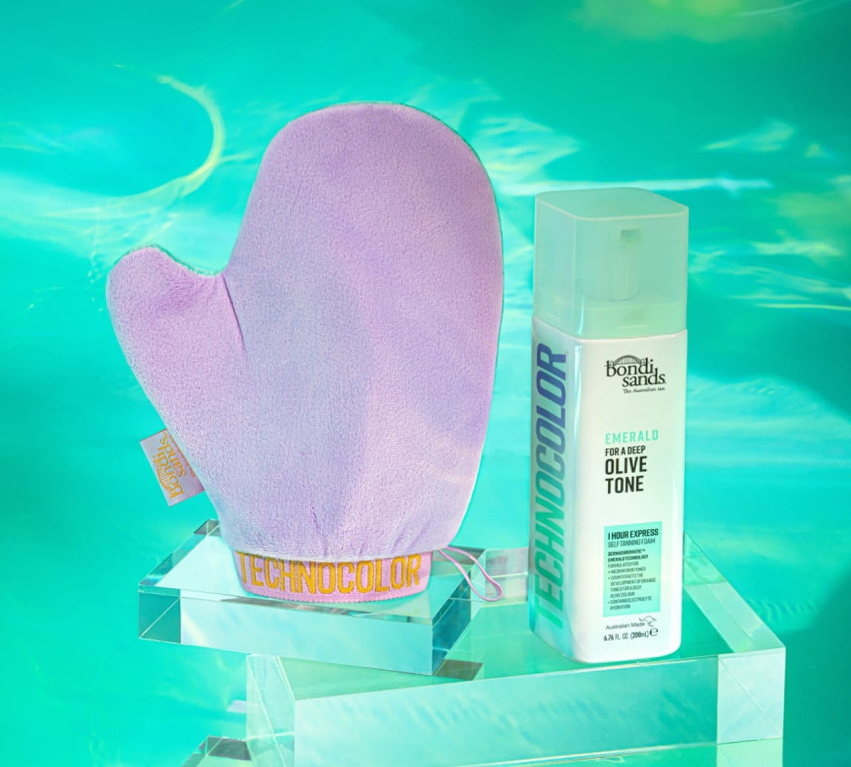 A purple tanning mitt and a bottle of BondiSandsEU Emerald Technocolor 1 Hour Express Self Tanning Foam for medium skin tones are shown on clear acrylic stands against a turquoise, water-inspired background.