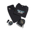 A flat lay of Bondi Babe Tanning PJs by BondiSandsEU—black shirt with “Bondi Babe”, black pants, self tanning sheet protector, small jar, makeup applicator, and black pouch—all on a white background.