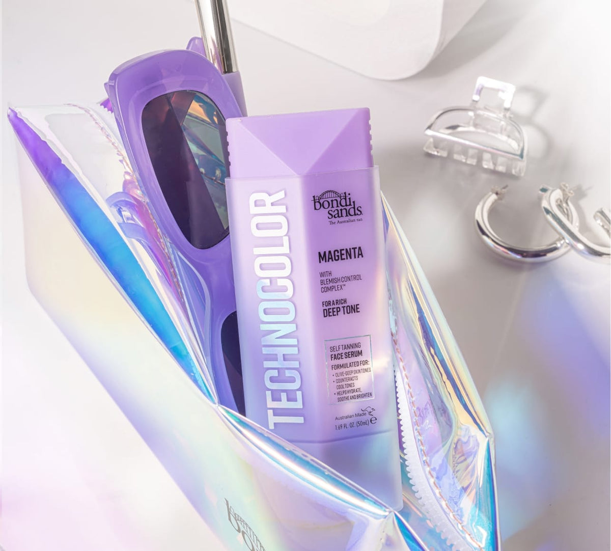 A holographic makeup bag holds purple sunglasses and a BondiSandsEU Magenta Technocolor Self Tanning Face Serum for olive-deep skin tones. Silver hoop earrings and a clear hair clip rest nearby on a white surface.