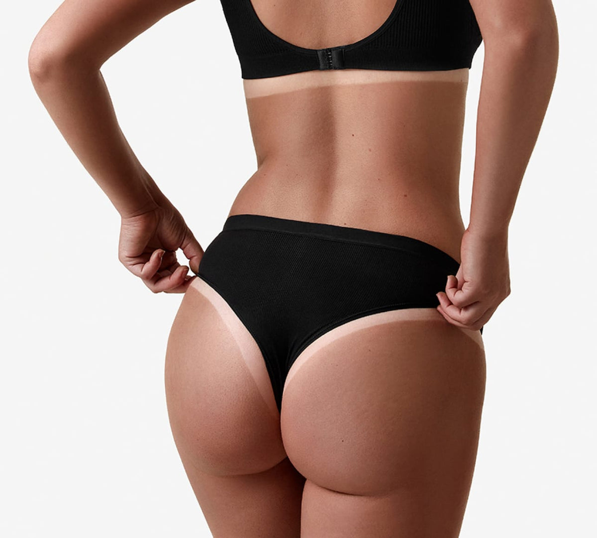 A person in black bra and underwear, seen from behind against a plain white background, adjusts the underwear with skin glowing as if enhanced by BondiSandsEU Salon Solution Light-Medium Professional Spray Tan.