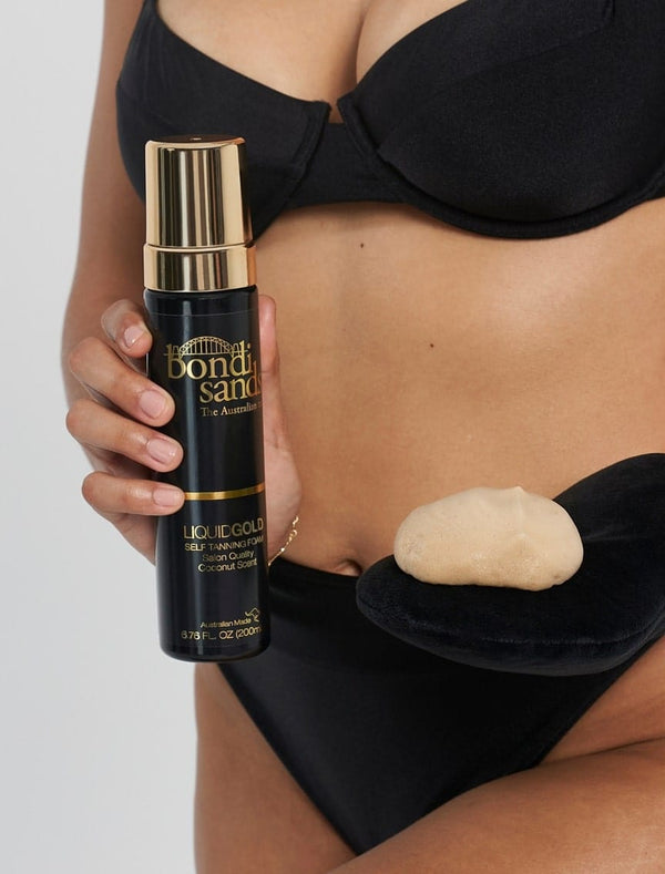 A person in a black bikini holds BondiSandsEU Self Tanning Foam Liquid Gold in one hand and a mitt with foam in the other, highlighting a radiant liquid gold glow against a plain background.