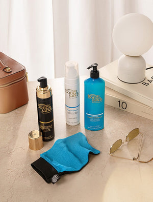 On a beige surface with books and a white curtain, the BondiSandsEU Self Tanning Foam Liquid Gold, two other skincare products, a brown bag, sunglasses, blue exfoliating mitt, and modern white lamp are displayed.