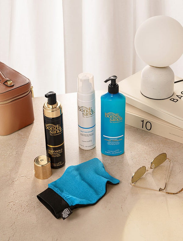 On a beige surface with books and a white curtain, the BondiSandsEU Self Tanning Foam Liquid Gold, two other skincare products, a brown bag, sunglasses, blue exfoliating mitt, and modern white lamp are displayed.