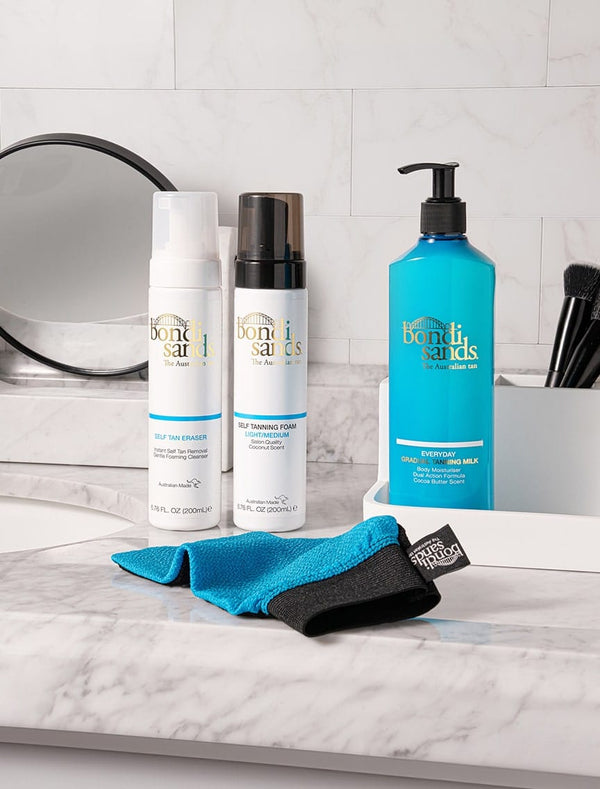 BondiSandsEU's Self Tanning Foam Light Medium and two other skincare products, plus a tanning mitt, are displayed on a marble bathroom counter with a mirror and makeup brushes in the background.