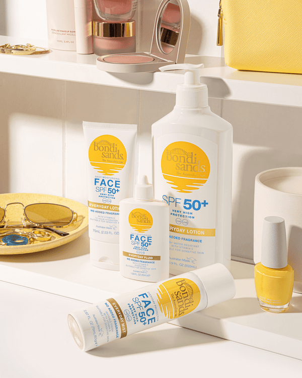 A BondiSandsEU SPF 50+ Everyday Sunscreen Lotion Pump sits on a yellow-themed bathroom counter, surrounded by sunglasses, a ring, nail polish, and a yellow purse for a sunny, stylish setup.