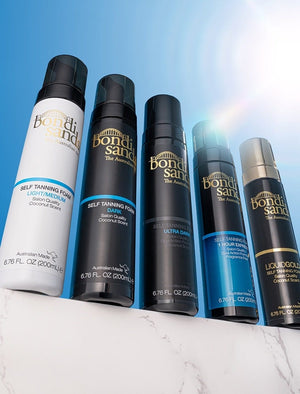 Five BondiSandsEU Self Tanning Foam Liquid Gold bottles stand upright on a white marble surface with a blue sky backdrop, showcasing a range of formulations from light/medium to ultra dark for every glow.