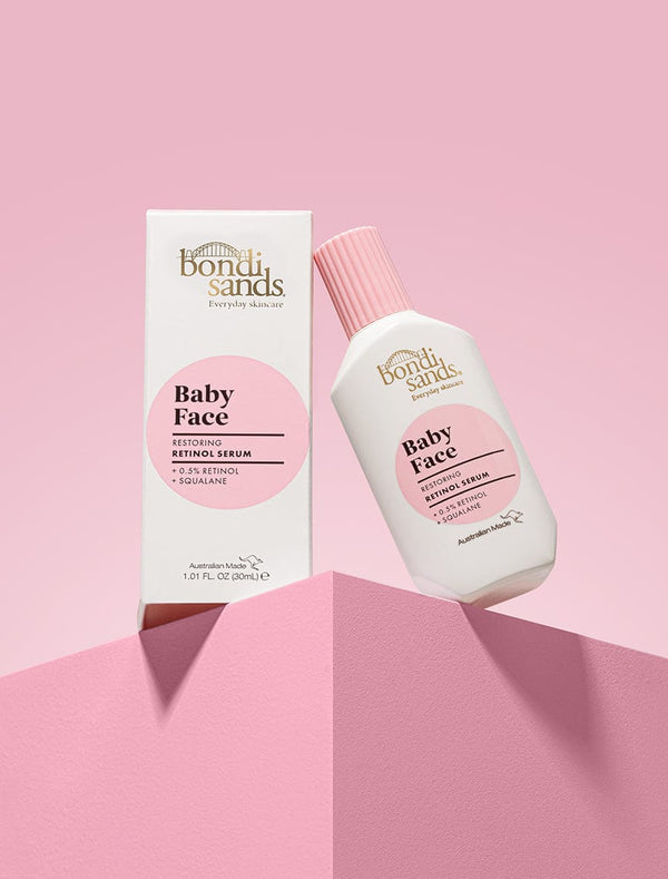 A white bottle and box of BondiSandsEU Baby Face Retinol Serum, with pink labels, are displayed on a pink geometric surface against a gradient pink background—ideal for reducing fine lines and supporting youthful-looking skin.