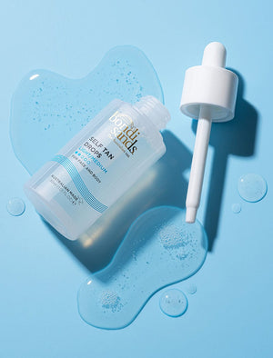A clear bottle of BondiSandsEU Self Tan Drops Light/Medium with a white dropper rests on a light blue surface, surrounded by tanning liquid and droplets, promising glowing skin and the perfect Australian tan.