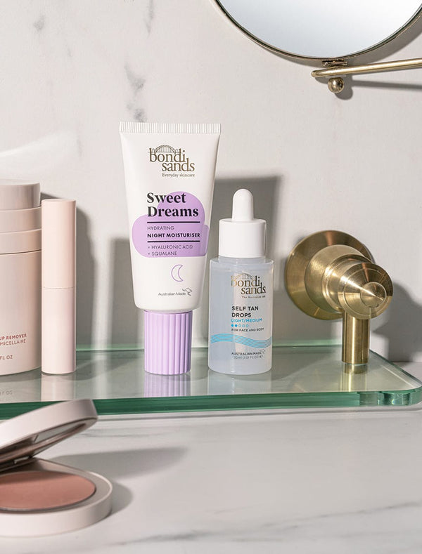 A glass bathroom shelf holds BondiSandsEU Self Tan Drops Light/Medium and Sweet Dreams night moisturizer among other beauty items, with a gold faucet and mirror in the background—get that flawless Australian tan from home.