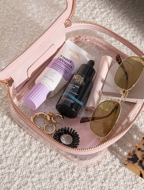 A clear pink zippered pouch on a carpet holds sunglasses, a small mirror, a black hair coil, gold hoop earrings, a tube labeled Sweet Dreams, BondiSandsEU Self Tan Drops Dark, and a pink lip product.