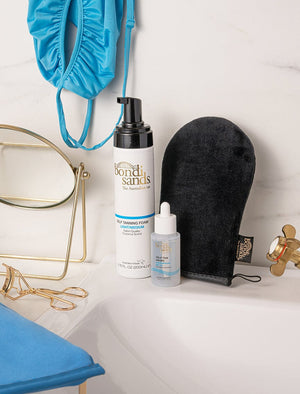 BondiSandsEU Self Tan Drops Light/Medium, a tanning mitt, and a blue swimsuit sit on a bathroom counter with a sink and gold faucet. Nearby, a vanity mirror and eyelash curler create an effortless Australian tan vibe.