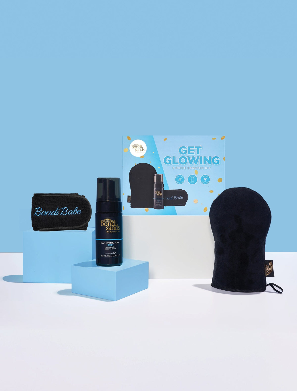The Bondi Sands Europe Dark Get Glowing 3 Piece Starter Kit—including a Self Tanning Foam, Application Mitt, and Bondi Babe headband—is showcased on blue and white platforms against a blue background for the ultimate self tan.