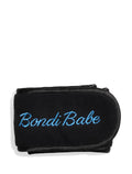 The Bondi Babe Headband by Bondi Sands Europe is a black terry cloth headband with "Bondi Babe" embroidered in light blue cursive, designed for hair protection. It features a velcro closure and appears against a white background.