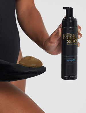 A person in a black swimsuit holds BondiSandsEU Self Tanning Foam Ultra Dark and a mitt with foam, ready to achieve the darkest tan, against a plain light background.