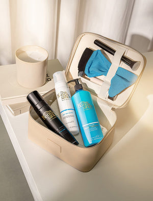 A beige cosmetic case on a white table holds three BondiSandsEU bottles, including Self Tanning Foam Ultra Dark for the deepest Australian tan, plus a tanning mitt and makeup brush. Nearby are a lit candle and closed book in sunlight.