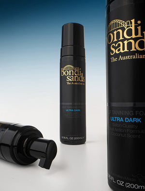 Three bottles of BondiSandsEU Self Tanning Foam Ultra Dark are arranged on a white surface with a blue gradient background, highlighting the sleek packaging and dispenser for your deepest Australian tan.