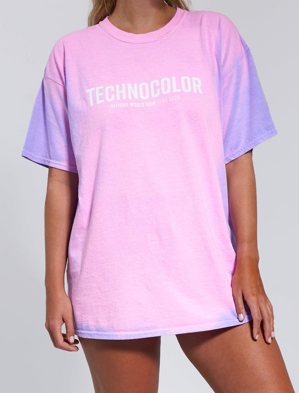 A person wears the Technocolor Technoverse Tee by BondiSandsEU—an oversized purple, short-sleeved shirt with "TECHNOCOLOR OFFICIAL WORLD TOUR APRIL 2023" printed in white across the chest, set against a plain light gray background.