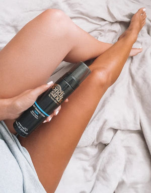 A person with tanned legs lies on a bed, holding a bottle from the BondiSandsEU Dark Foam Bundle. One leg is visibly more tanned than the other, showing the before and after effect of this must-have self-tanning product.